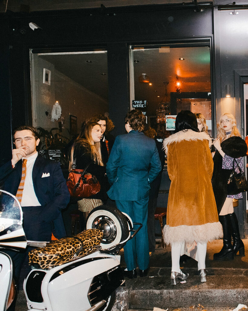 A stylish group of people gathers outside a dimly lit café at night; one Tickety Boo man in a suit stands near a white scooter with a leopard print seat, while others chat by the entrance, dressed in fashionable retro clothing.