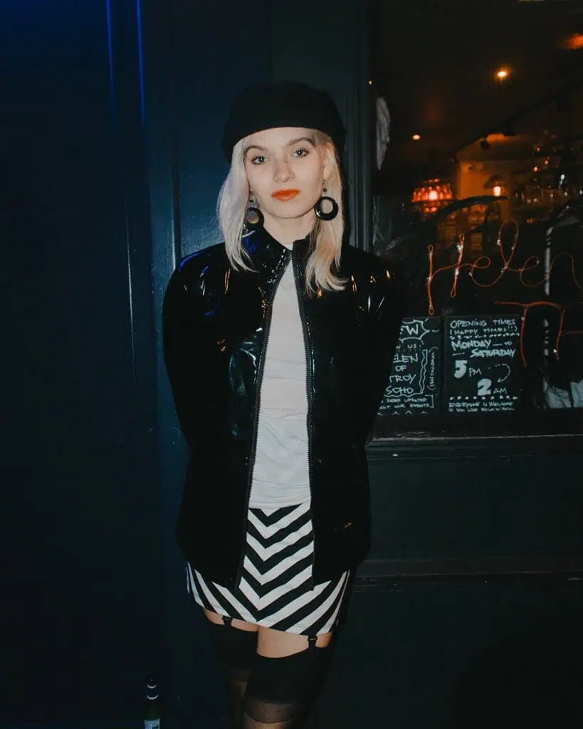 A woman with blonde hair stands against a dark wall wearing a black hat, black jacket, white shirt, and a Tickety Boo black-and-white zigzag skirt. She has large earrings and is looking directly at the camera.