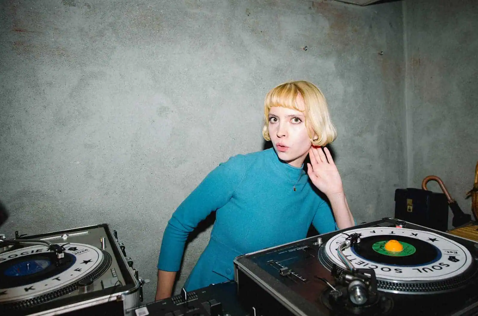 A person with short blonde hair and a blue dress stands between two turntables, one hand to their ear as if listening closely. The plain, gray concrete walls add to the Tickety Boo vibe of this DJ scene.