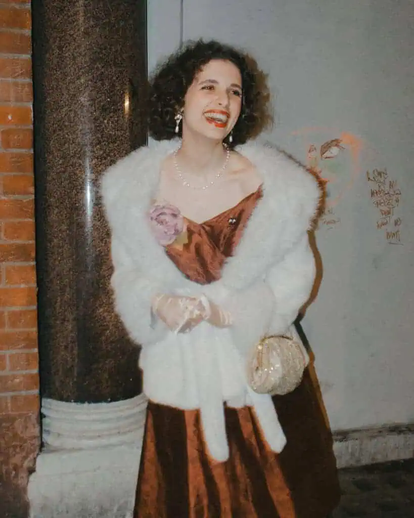 A woman in a brown satin dress and white fur coat smiles brightly in a tickety boo mood. She wears pearl jewelry, holds a gold purse, and stands by a brick wall with graffiti in the background.