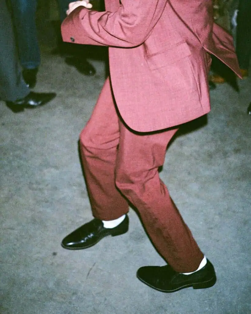 A person wearing a red suit, white socks, and black dress shoes is captured mid-dance on a concrete floor, looking absolutely tickety boo. The photo shows only their body from the shoulders down, with other people partially visible in the background.