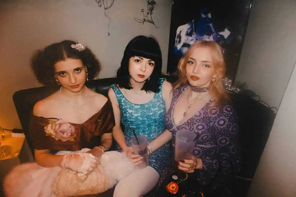 Three women in vintage-style dresses sit closely together on a dark sofa at a Tickety Boo party, each holding a drink. The background is dimly lit, with a blurred poster or painting on the wall behind them.