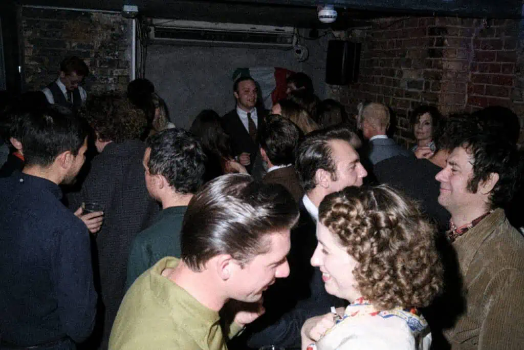 A crowded bar or club with people socializing, talking, and smiling in dim lighting. Exposed brick walls and a visible air conditioner set the scene as guests in vintage or retro-style clothing enjoy a tickety boo night out.