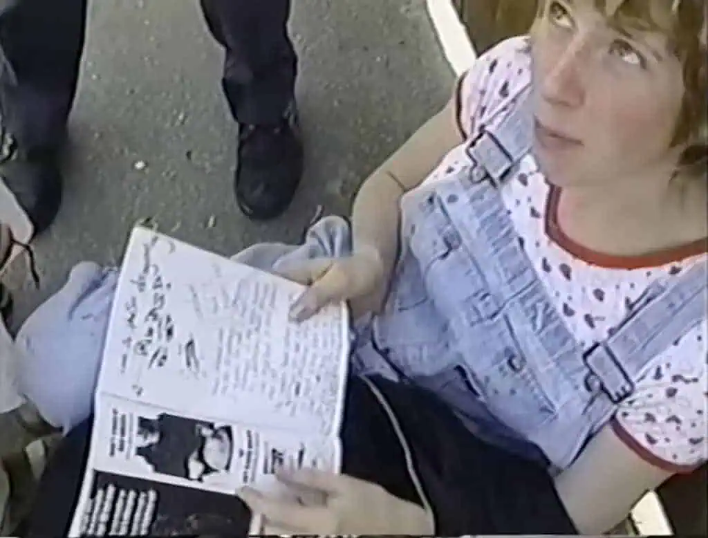 A person in a light blue denim overall dress and a white shirt with red patterns sits outdoors, holding open a notebook with handwritten notes and black-and-white printed images, reminiscent of scenes from the Dirty Girls documentary. Other people’s legs are visible nearby.