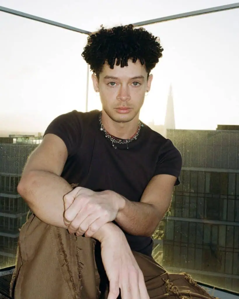 A person with curly hair sits on a rooftop at sunset, channeling Erykah Badu vibes in a black t-shirt, brown pants, and a chain necklace, with modern buildings and a glass railing in the background.