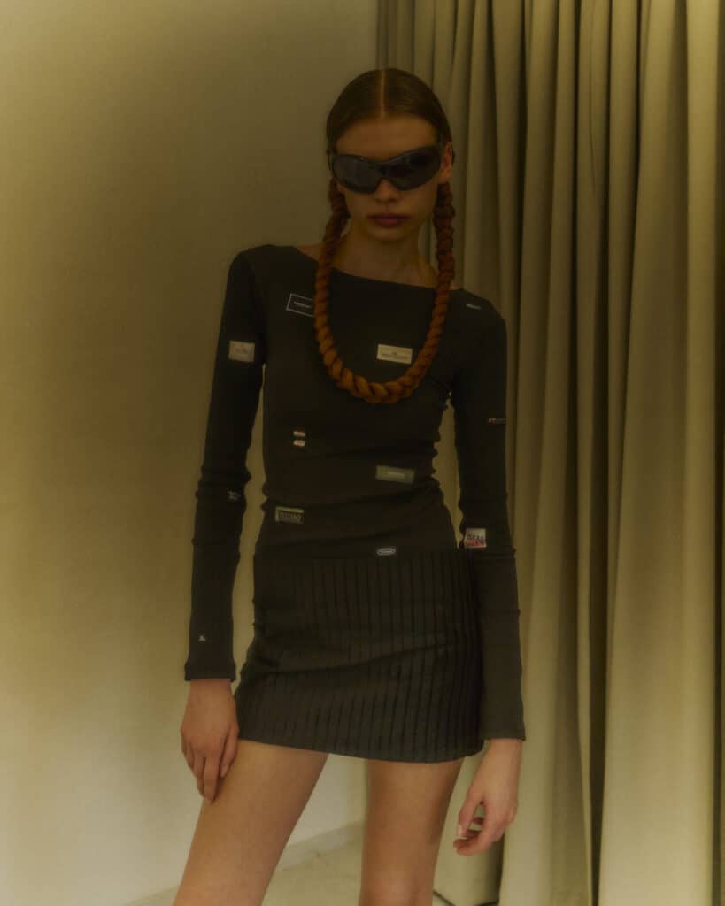 A woman with braided hair wears dark sunglasses, a long-sleeve black top with small labels, and a black pleated mini skirt, standing indoors in front of beige curtains.