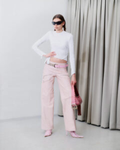 A person poses indoors wearing a fitted white long-sleeve top, light pink wide-leg pants with a pink belt, pink patterned boots, large black sunglasses, and carries a pink studded handbag. Beige curtains hang in the background.
