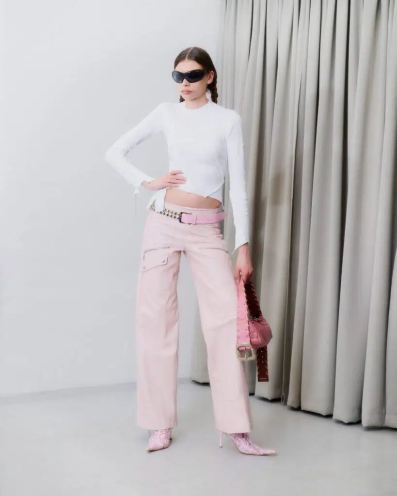 A person poses indoors wearing a fitted white long-sleeve top, light pink wide-leg pants with a pink belt, pink patterned boots, large black sunglasses, and carries a pink studded handbag. Beige curtains hang in the background.