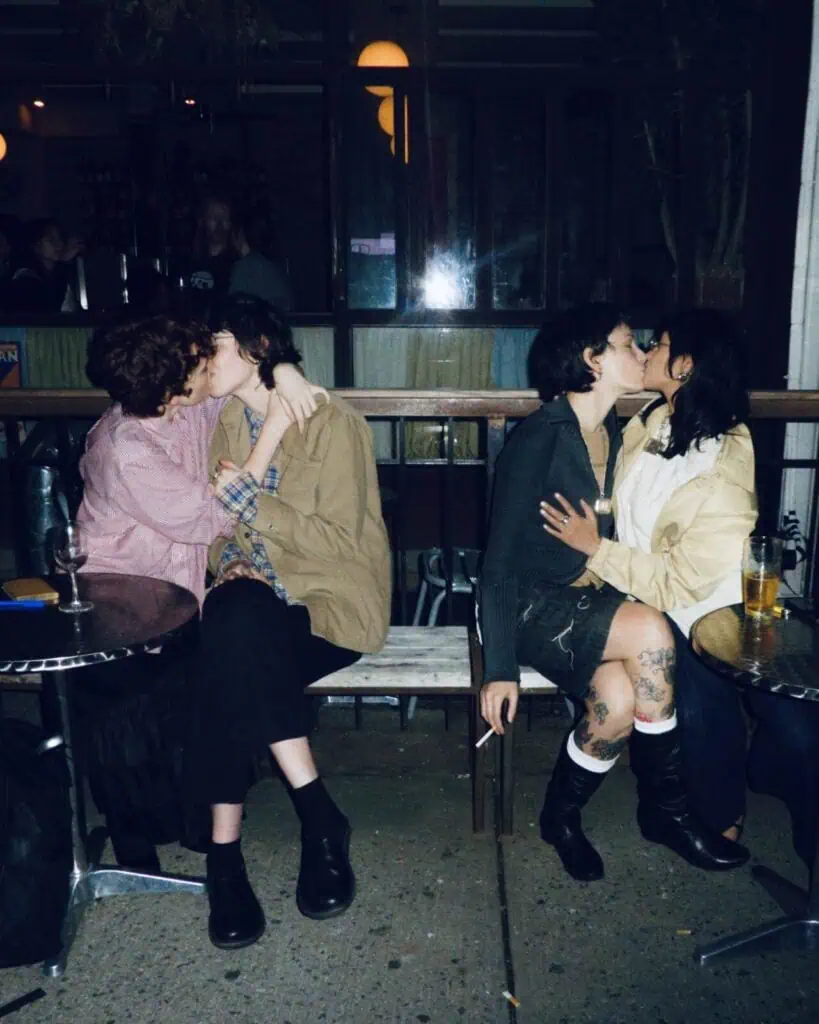 Two couples sit closely together on a bench outside a bar at night, each pair kissing. There are drinks on the tables beside them, and the scene has a warm, intimate, and candid atmosphere.