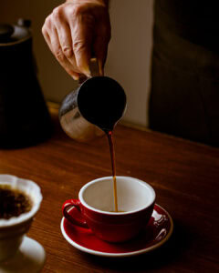 A hand pours Aliena Coffee from a metal pitcher into a red cup and saucer on a wooden table, with a filter and coffee grounds visible in the lower left corner.