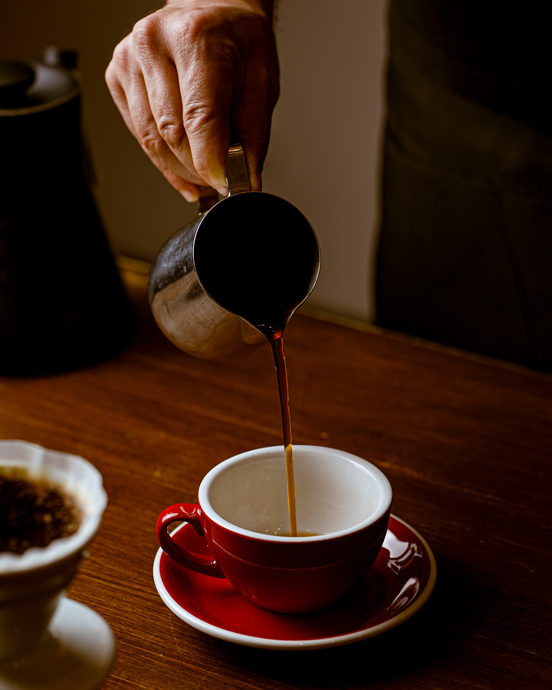 A hand pours Aliena Coffee from a metal pitcher into a red cup and saucer on a wooden table, with a filter and coffee grounds visible in the lower left corner.