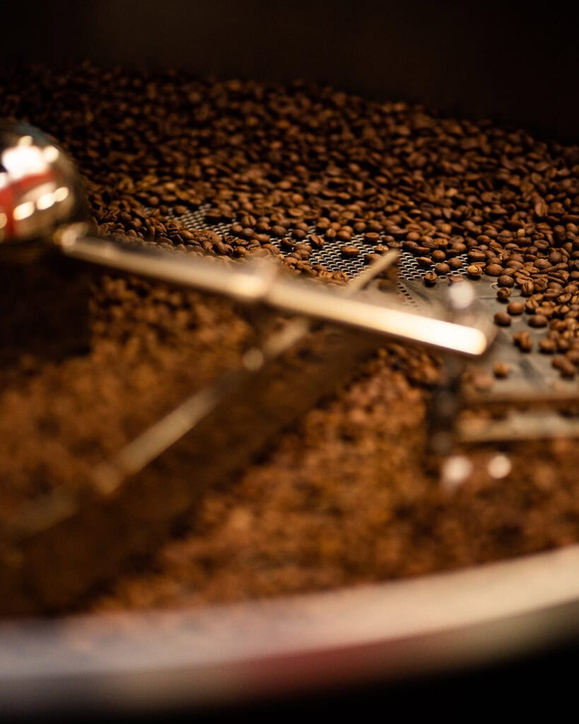 A close-up of roasted Aliena coffee beans being stirred by metal arms inside a coffee roasting machine. The image focuses on the beans, showing their rich brown color and glossy texture.