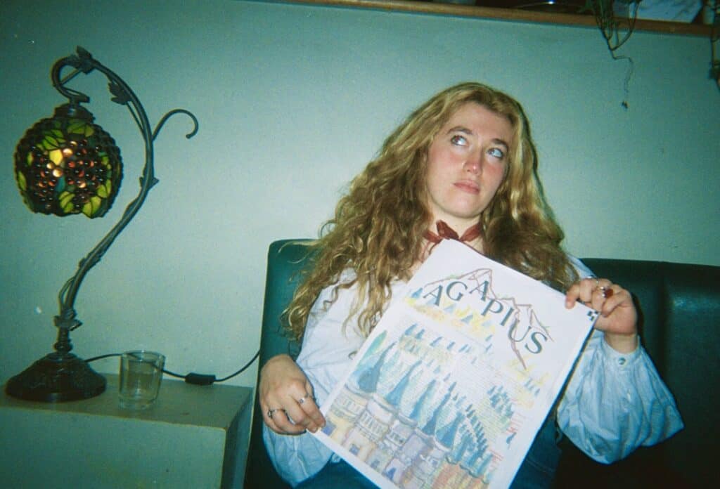 A woman with long curly hair sits on a green bench, holding a colorful illustrated board game box labeled AGAPUS. A stained glass lamp and a glass of water are on a nearby table. She looks upward thoughtfully.