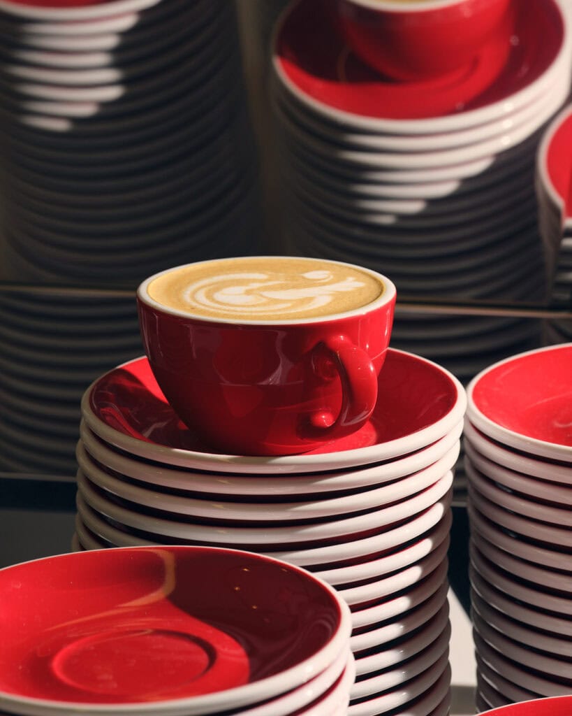 Aliena red coffee with latte art sits on a stack of red and white saucers, surrounded by more stacked saucers in the background.
