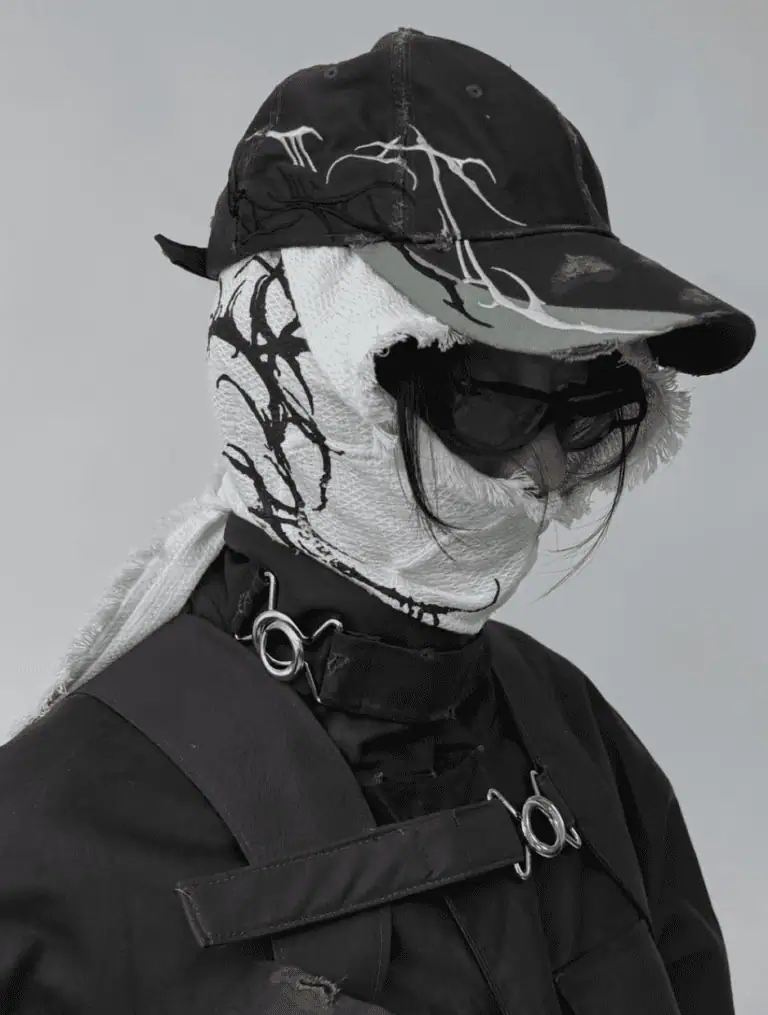 A person wearing a dark cap and sunglasses, with a white FFFPOSTAL scarf featuring black abstract patterns wrapped around their head and neck. They are dressed in a dark jacket with metal rings and straps.