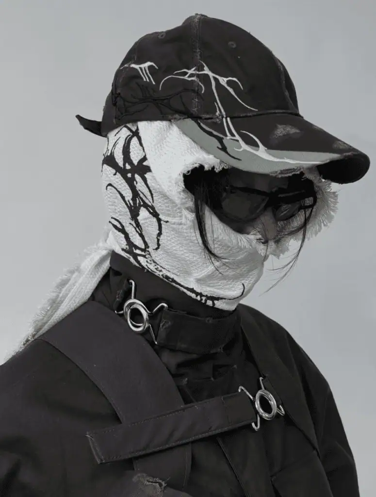 A person wearing a dark cap and sunglasses, with a white FFFPOSTAL scarf featuring black abstract patterns wrapped around their head and neck. They are dressed in a dark jacket with metal rings and straps.