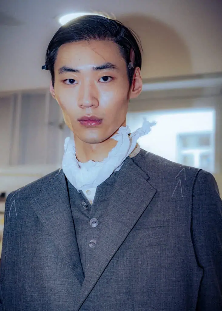 A person with short dark hair wears a grey Sonia Carrasco three-piece suit and a white scarf around their neck. They look directly at the camera in a well-lit indoor setting.