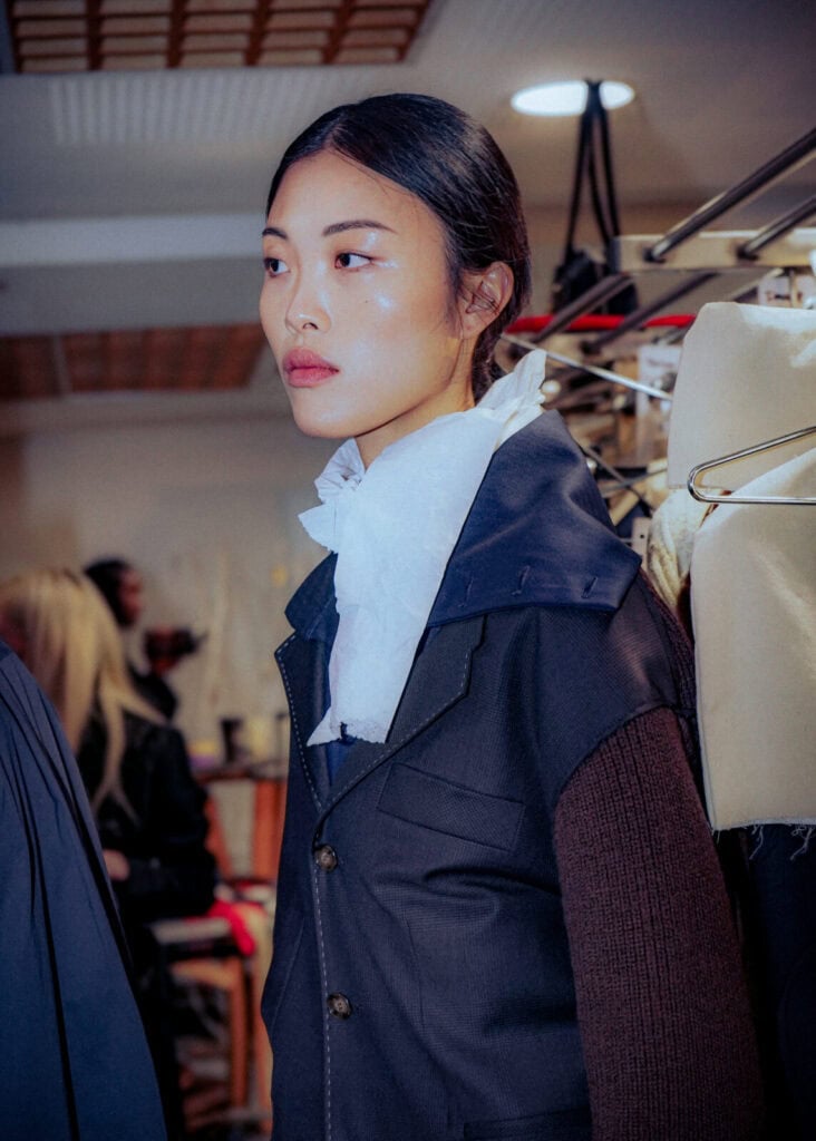 A woman with straight dark hair tied back stands indoors wearing a navy jacket and a large white ruffled scarf. She looks to the side, with racks of clothes and people blurred in the background.