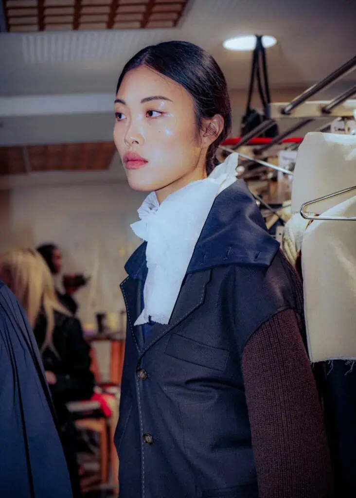 A woman with straight dark hair tied back stands indoors wearing a Sonia Carrasco navy jacket and a large white ruffled scarf. She looks to the side, with racks of clothes and people blurred in the background.