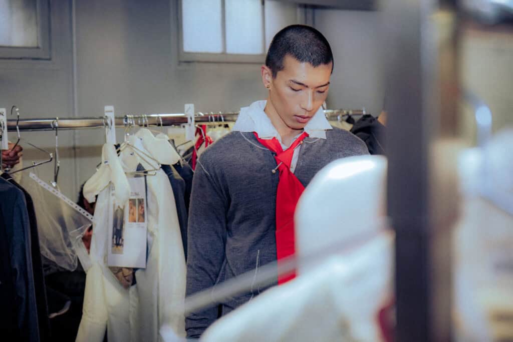 A young man with short hair stands in front of a clothing rack, wearing a gray hoodie over a white shirt and red tie, looking down in a room with hanging clothes and soft natural light.