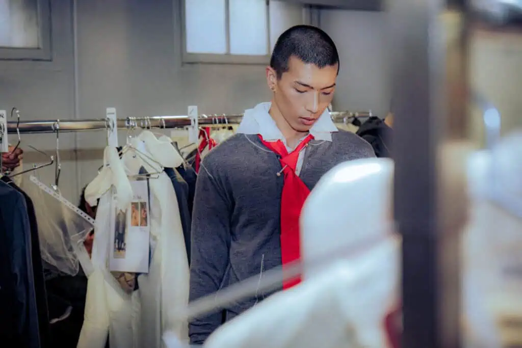 A young man with short hair stands in front of a clothing rack, wearing a gray hoodie over a white shirt and red tie, looking down in a room with hanging clothes and soft natural light at the Sonia Carrasco backstage.