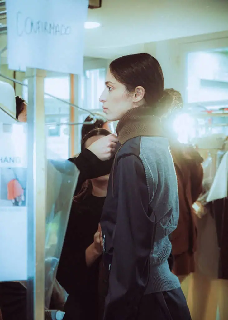 A woman stands in profile backstage as a person adjusts her clothing. She wears a dark Sonia Carrasco outfit and her hair is tied back. There are racks of clothes and a sign that reads Confirmado in the background.