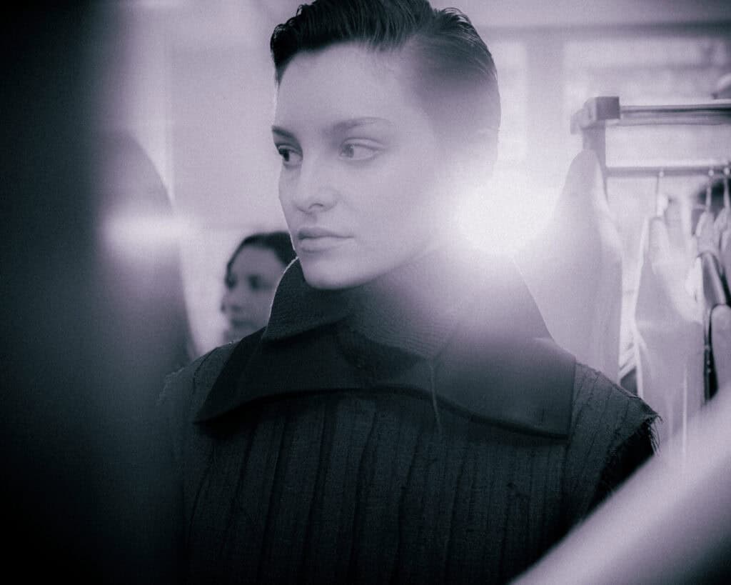 A woman with short hair and a high-collared dark outfit stands indoors, looking to the side. The background is softly blurred with bright light shining behind her.