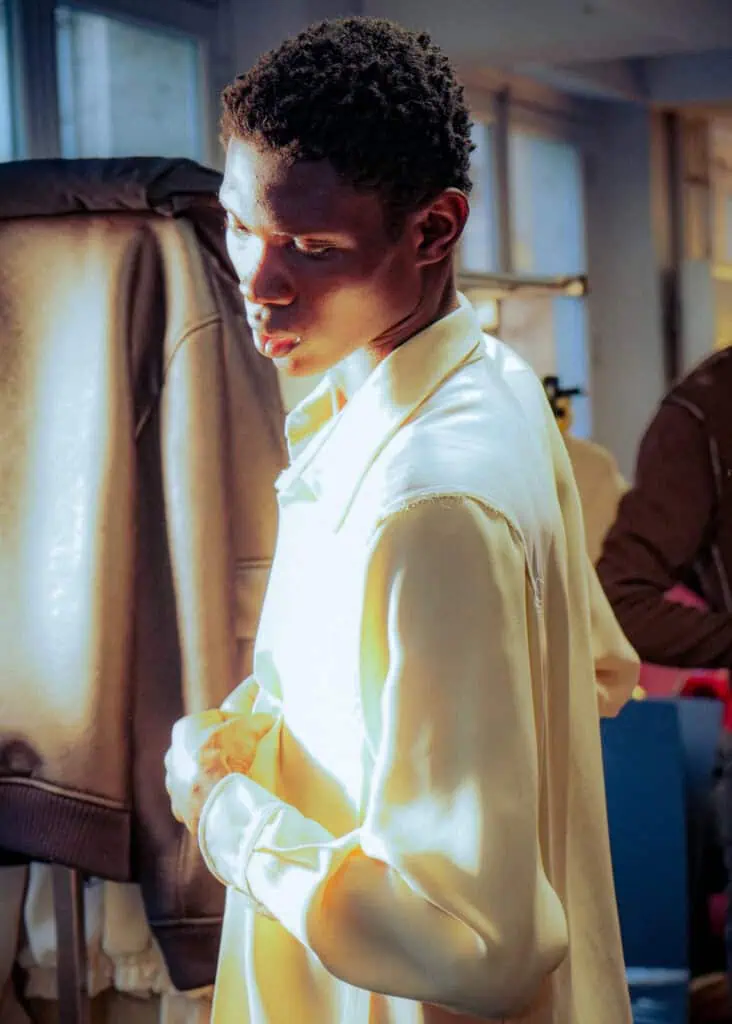 A person in a light-colored, silky shirt stands indoors, bathed in soft, natural light. They are looking slightly down and to the side while buttoning their shirt, with a jacket and blurred figure in the background at the Sonia Carrasco backstage.