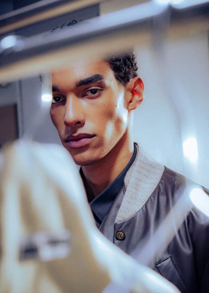 A young man with short curly hair and sharp features gazes intently at the camera, partially obscured by an out-of-focus hanger and clothing in the foreground. The lighting is soft and highlights his face.