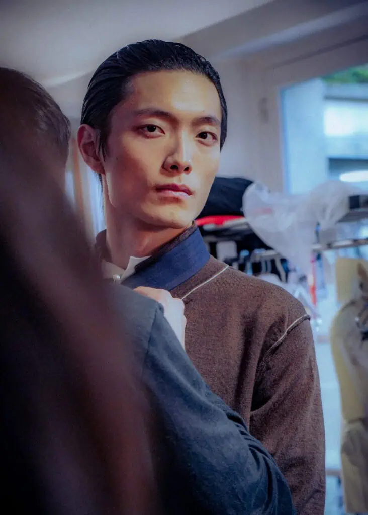 A young man with slicked-back hair looks into the camera while someone adjusts the collar of his dark jacket. He stands indoors near a window, with blurred clothing and hangers in the background at the Sonia Carrasco backstage.