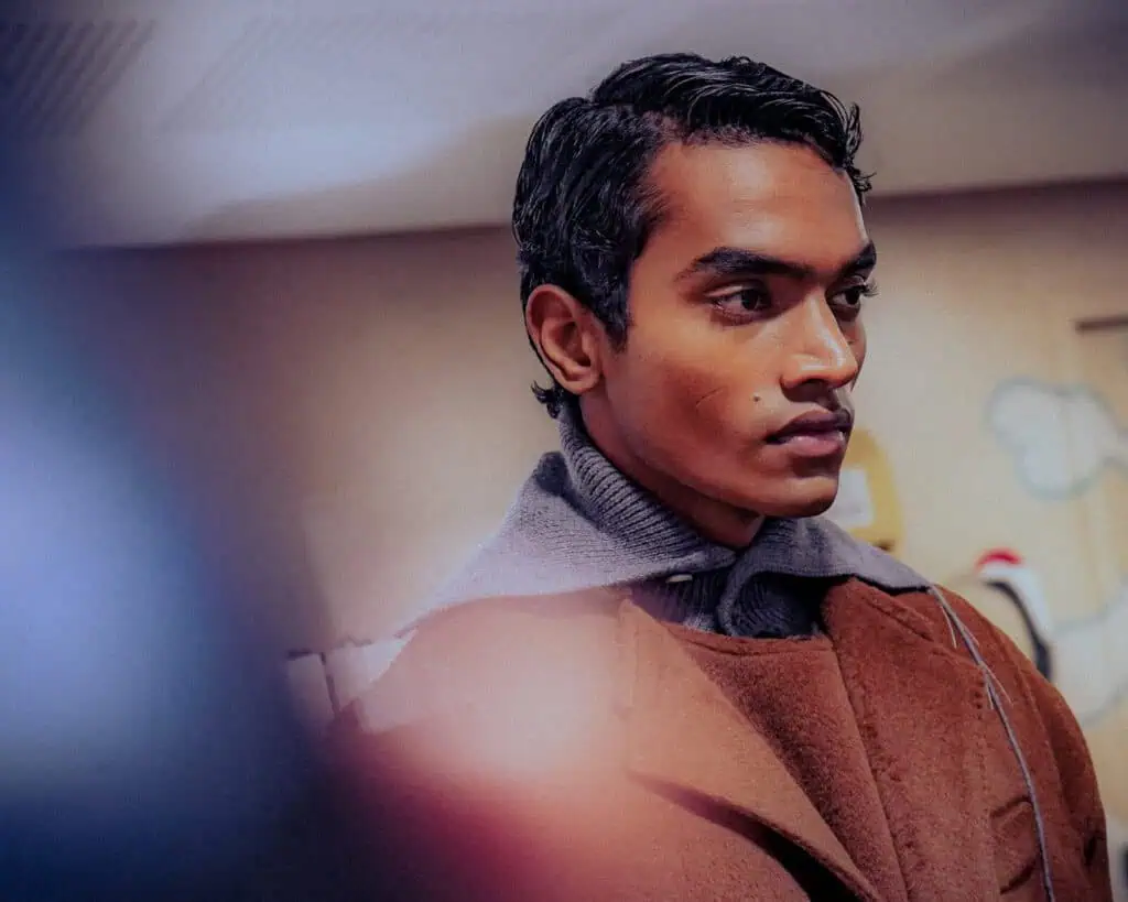 A man with styled dark hair wears a brown wool coat layered over a gray turtleneck, looking serious and gazing to the side in an indoor setting with soft lighting and a blurred background.