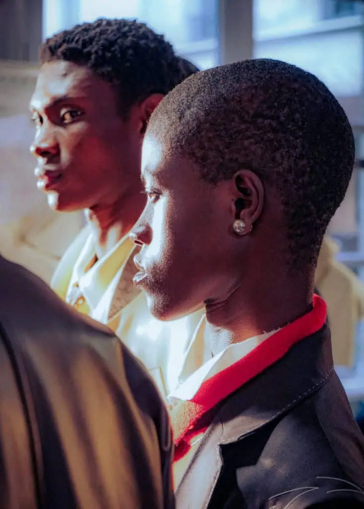 Two men in formal attire stand side by side, one in profile wearing a dark blazer and red shirt with an earring, and the other slightly blurred in the background wearing a light-colored jacket. They are indoors with natural light.