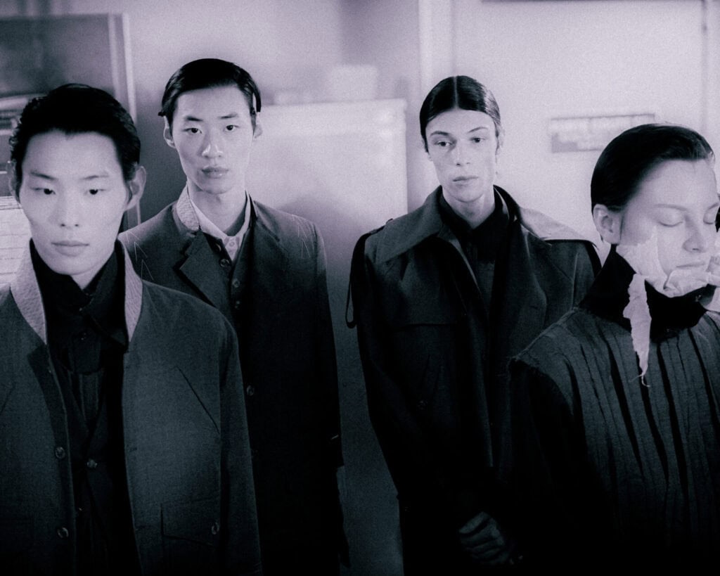 Four individuals stand indoors wearing dark, structured clothing. Three look away with neutral expressions, while one has their eyes closed and face partially covered with white tape. The scene has a somber, muted tone.