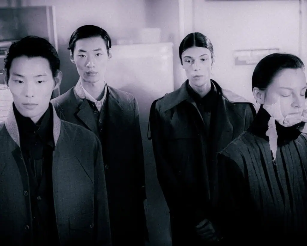 Four individuals stand indoors wearing dark, structured Sonia Carrasco clothing. Three look away with neutral expressions, while one has their eyes closed and face partially covered with white tape. The scene has a somber, muted tone.