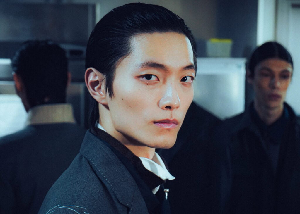 A person with slicked-back black hair and a serious expression wears a dark suit and stands indoors, facing the camera. Two other people, also dressed formally, are blurred in the background.