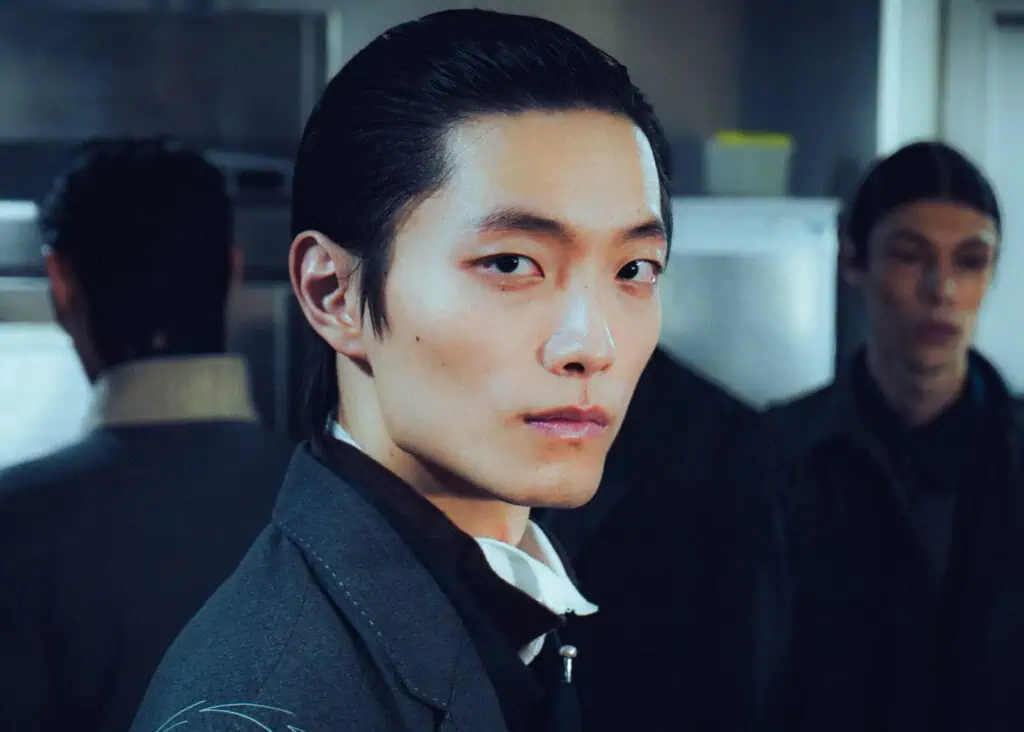A person with slicked-back black hair and a serious expression wears a dark suit and stands indoors, facing the camera. Two other people, also dressed formally, are blurred in the background.