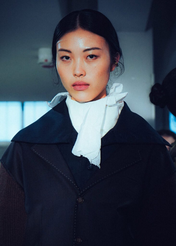 A woman with straight black hair wearing a dark outfit, a white neck scarf, and a cape-like collar stands indoors, looking directly at the camera under soft lighting.
