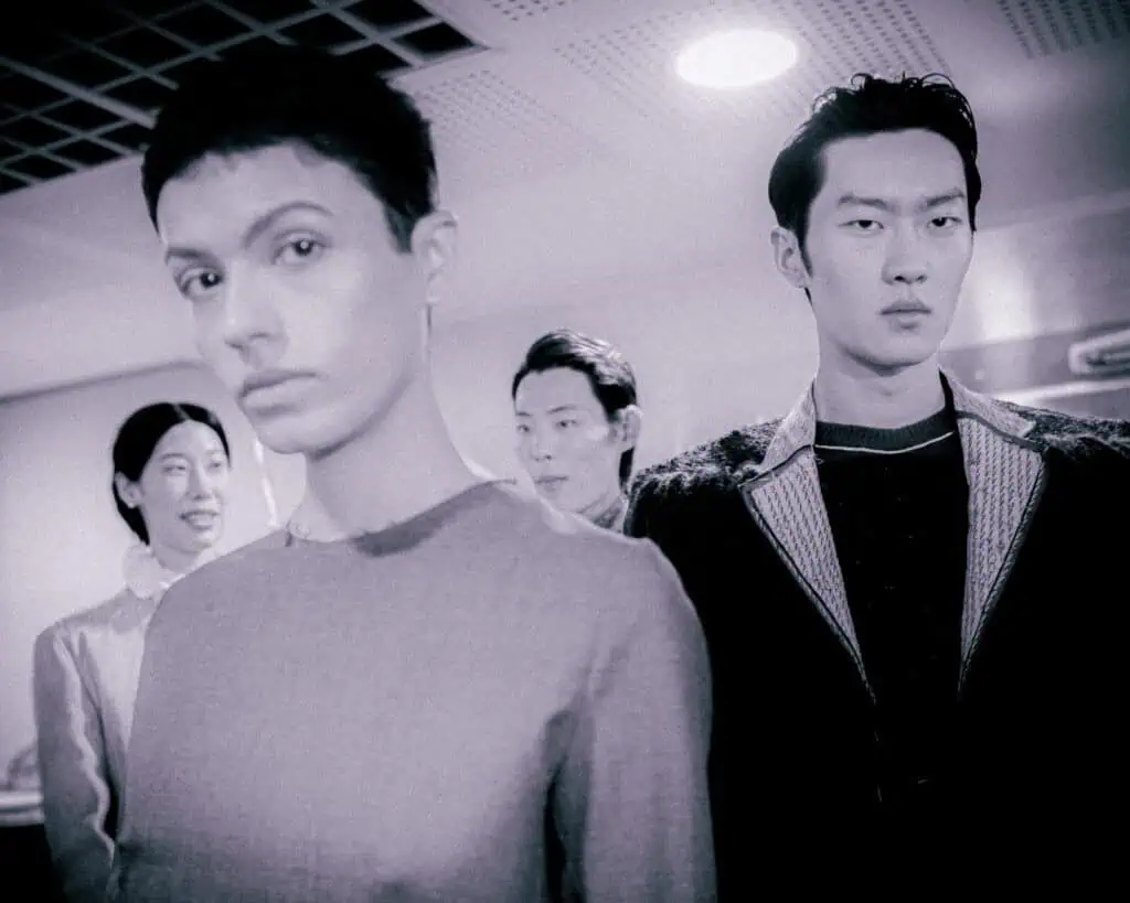 A group of four people stand indoors under bright ceiling lights, with two individuals in the foreground looking serious and two others behind them, slightly out of focus. The image has a muted, monochrome tone.