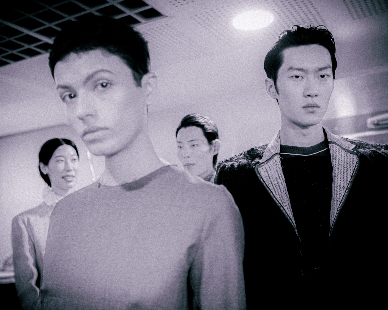 A group of four people stand indoors under bright ceiling lights, with two individuals in the foreground looking serious and two others behind them, slightly out of focus. The image has a muted, monochrome tone.