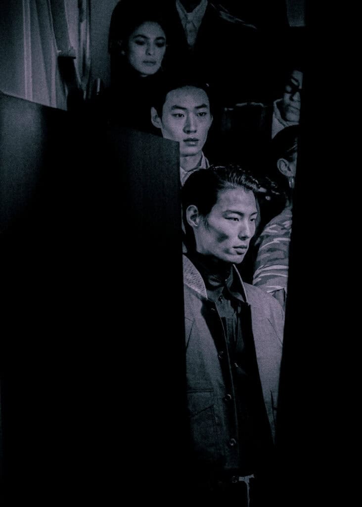 A group of people, mostly men, stand in a dimly lit space. Dramatic shadows and dark panels partially obscure their faces, creating a mysterious and moody atmosphere.