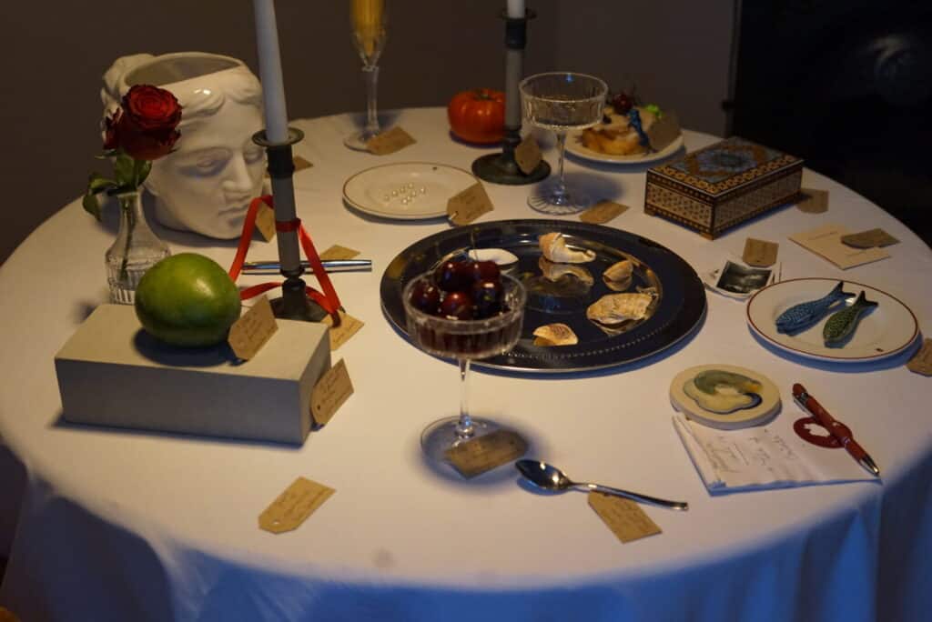 A round table set with eclectic items, including a fruit bowl, candles, a classical head vase with a red rose, a mango, a goblet of cherries, a tomato, plates with food, and various handwritten labels next to the objects.