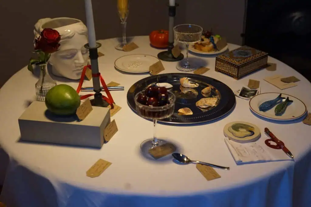 A round table set with eclectic items, including a fruit bowl, candles, a classical head vase with a red rose, a mango, a goblet of cherries, a tomato, plates with food, and various handwritten labels next to the objects.