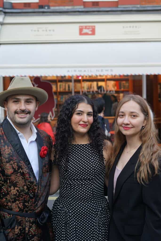 Diego Fernandez and Aryana Assl pose and smile outdoors in front of a bookstore for the Perediza IV presentation. The person on the left wears a hat and a patterned jacket, the center person wears a black and white polka dot dress, and the right person wears a black blazer.
