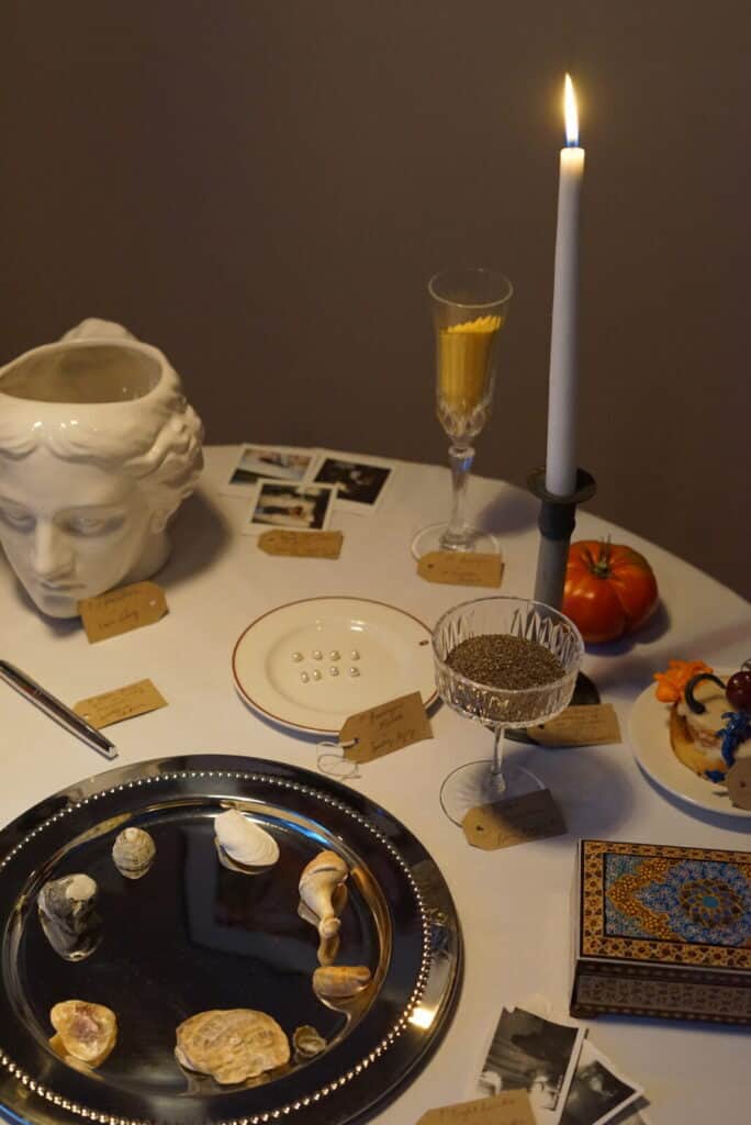 A round table at Perediza IV presentation displays seashells, a silver tray, a candle, a statue head, vintage photos, a plate with pills, labeled tags, a crystal glass with seeds, a tomato, and a slice of cake with flower decorations.