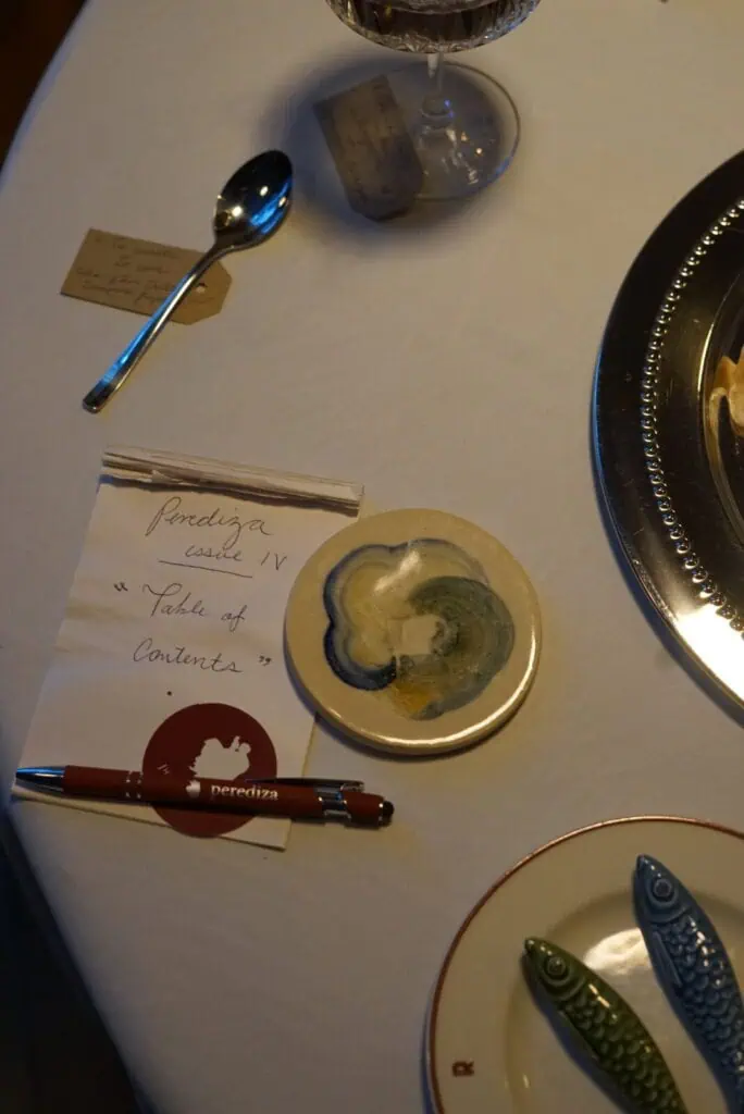 A notepad labeled Porediza Castle IV Table of Contents sits on a table with a red pen, a round ceramic coaster, a spoon, a glass of wine, and fish-shaped chopstick rests on a white tablecloth.