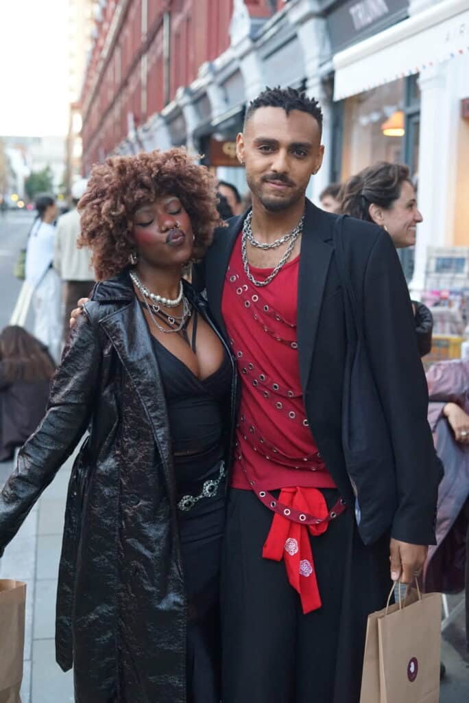 Two stylish people pose together on a city street during the Perediza IV presentation. The person on the left has curly hair, wears a shiny black coat, and layered necklaces. The person on the right wears a red shirt with silver details and holds a shopping bag.