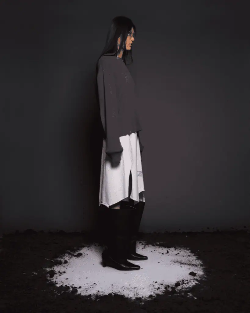 A woman stands in profile on a dark stage, wearing a loose purple sweater, a white skirt with a side slit, and black knee-high boots, surrounded by a circular patch of white powder on the ground.