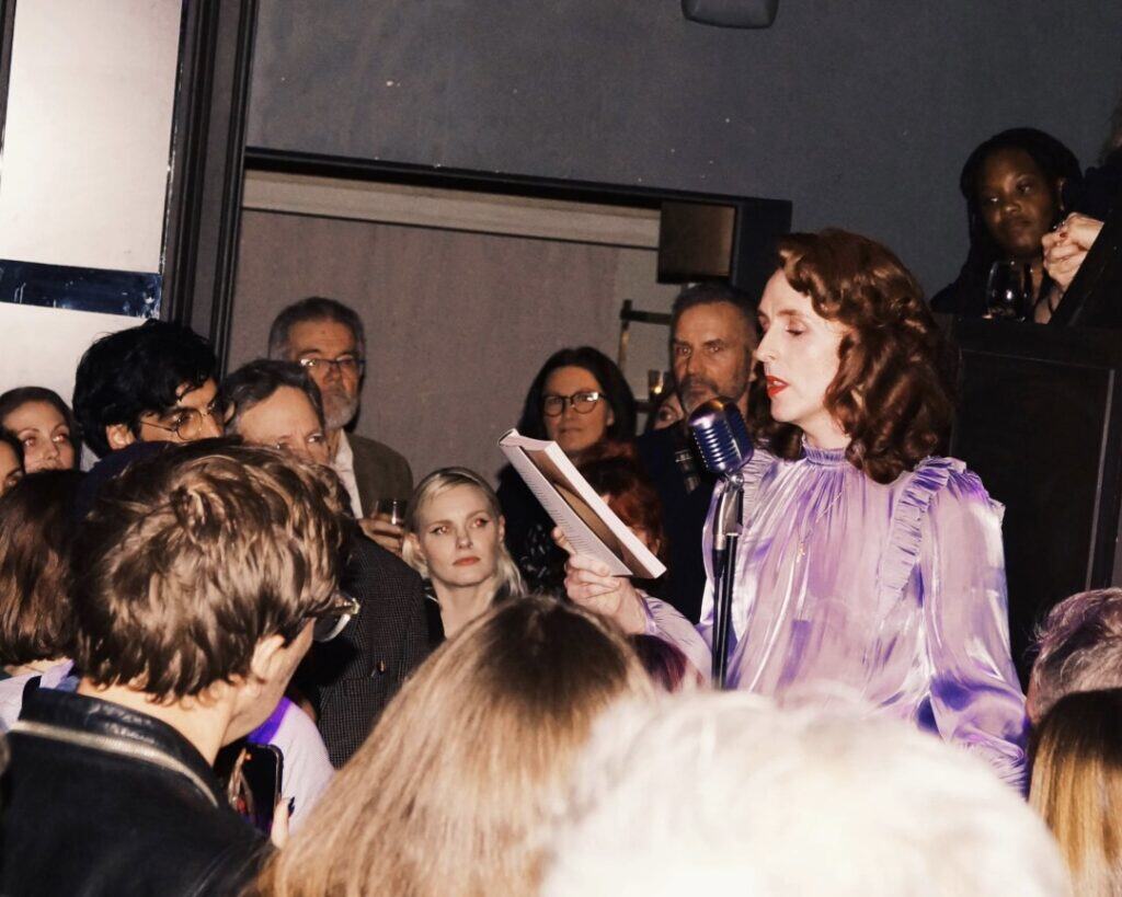 A woman with red hair in a shiny lavender blouse reads from a paper into a vintage microphone, surrounded by a diverse crowd in a dimly lit room, all attentively watching her.