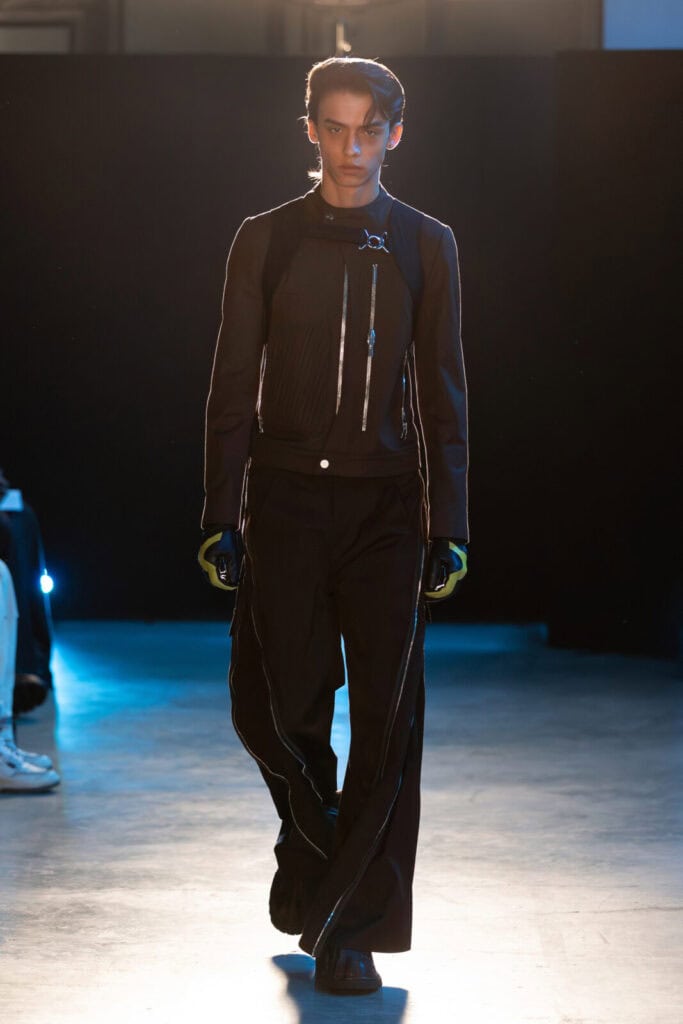 A model walks a runway wearing a black zip-up FFFPOSTAL jacket with metallic accents, wide-leg black pants with zippers, black gloves, and chunky black shoes, against a dark background with blue lighting.