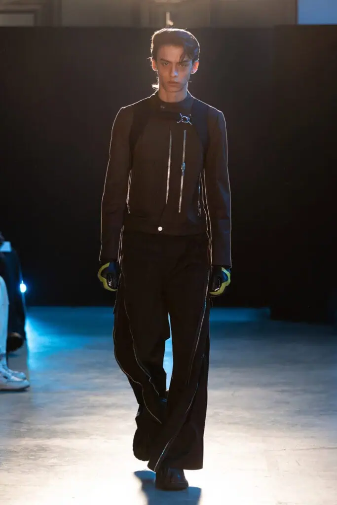 A model walks a runway wearing a black zip-up FFFPOSTAL jacket with metallic accents, wide-leg black pants with zippers, black gloves, and chunky black shoes, against a dark background with blue lighting.